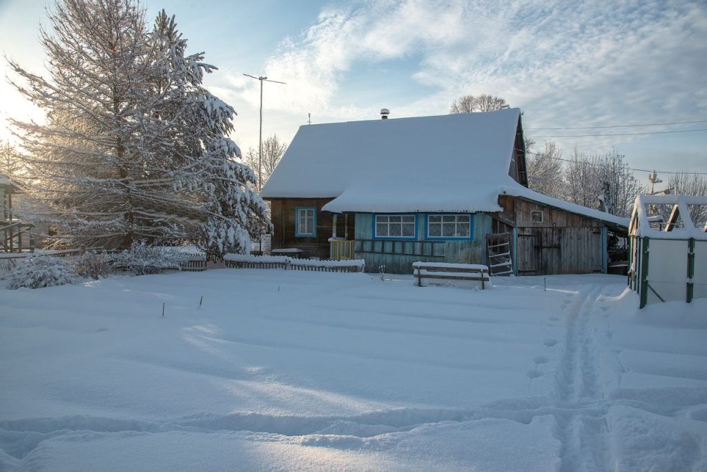 A picturesque wooden house blanketed in snow, surrounded by a serene winter landscape and clear blue sky.